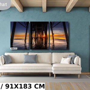 May include: A three-panel canvas print of a sunset over the ocean with a pier in the foreground. The print is 36 inches by 72 inches and is displayed on a white couch in a living room.