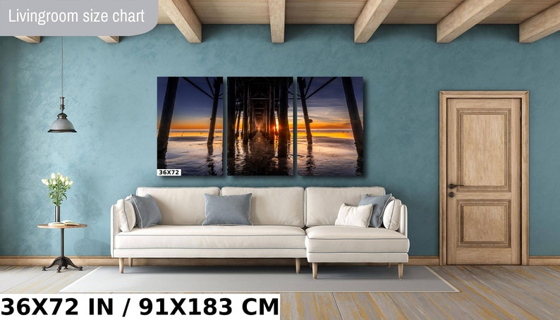 May include: A three-panel canvas print of a sunset over the ocean with a pier in the foreground. The print is 36 inches by 72 inches and is displayed on a white couch in a living room.