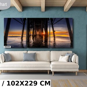 May include: A three-panel canvas print depicting a sunset over a wooden pier with the text "40X90" in the bottom left corner. The print is displayed on a white couch in a living room setting. The text "40X90 IN / 102X229 CM" is displayed below the couch.