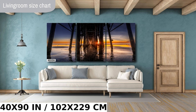 May include: A three-panel canvas print depicting a sunset over a wooden pier with the text "40X90" in the bottom left corner. The print is displayed on a white couch in a living room setting. The text "40X90 IN / 102X229 CM" is displayed below the couch.