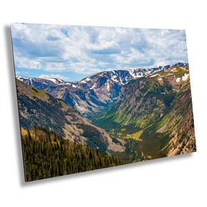 May include: A panoramic view of a mountain valley with snow-capped peaks, lush green slopes, and a winding road in the distance. The sky is blue with white clouds.