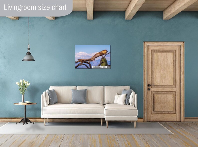 May include: A framed canvas print of a bird perched on a branch, set against a blue sky with clouds. The print is labeled with dimensions of 24x36 inches / 61x91 cm. The artwork hangs above a cream-colored sofa.