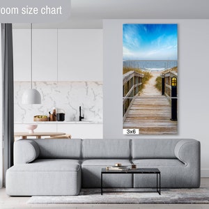 May include: A 3x6 foot canvas print of a wooden walkway leading to a beach with blue sky and ocean in the background. The walkway has a wooden railing and a lamp post on each side.