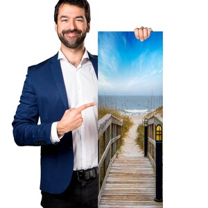 May include: A man in a blue suit points to a 1x3 photograph of a wooden walkway leading to a beach with blue sky and ocean in the background.