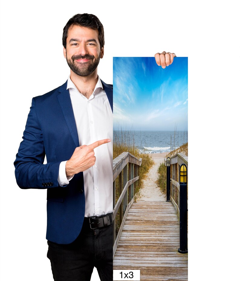 May include: A man in a blue suit points to a 1x3 photograph of a wooden walkway leading to a beach with blue sky and ocean in the background.