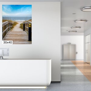 May include: A photograph of a wooden walkway leading to a beach. The walkway is lined with wooden railings and has a lamp post at the end. The beach is sandy and has blue water in the background. The image is 76.2 cm by 101.6 cm.