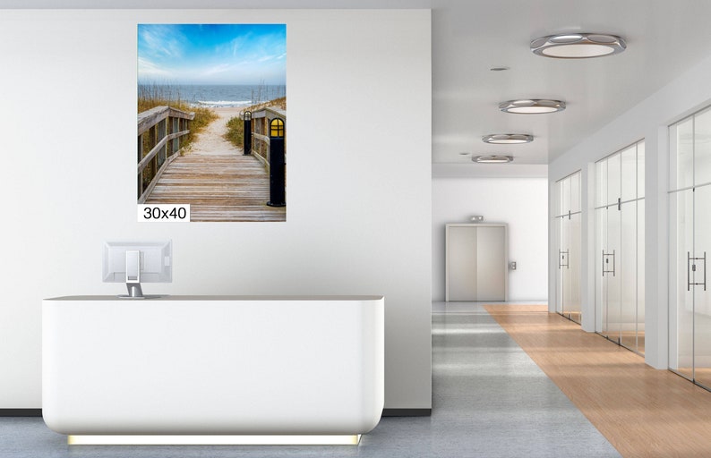 May include: A photograph of a wooden walkway leading to a beach. The walkway is lined with wooden railings and has a lamp post at the end. The beach is sandy and has blue water in the background. The image is 76.2 cm by 101.6 cm.