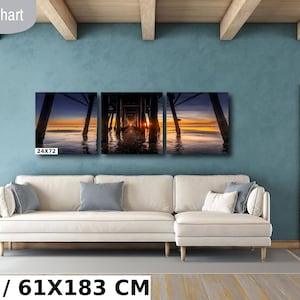 May include: A three-piece canvas print set depicting a sunset view under a pier. The print is displayed on a white couch in a living room setting. The print measures 24x72 inches or 61x183 centimeters.