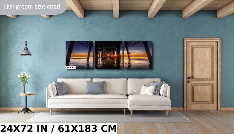 May include: A three-piece canvas print set depicting a sunset view under a pier. The print is displayed on a white couch in a living room setting. The print measures 24x72 inches or 61x183 centimeters.