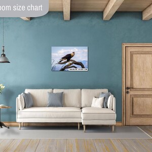 Op de afbeelding: Een ingelijste print van een roofvogel die op een tak zit, tegen een blauwe lucht. Het kunstwerk wordt tentoongesteld boven een cr&egrave;mekleurige bank in een woonkamer. De print meet 61x91 cm.