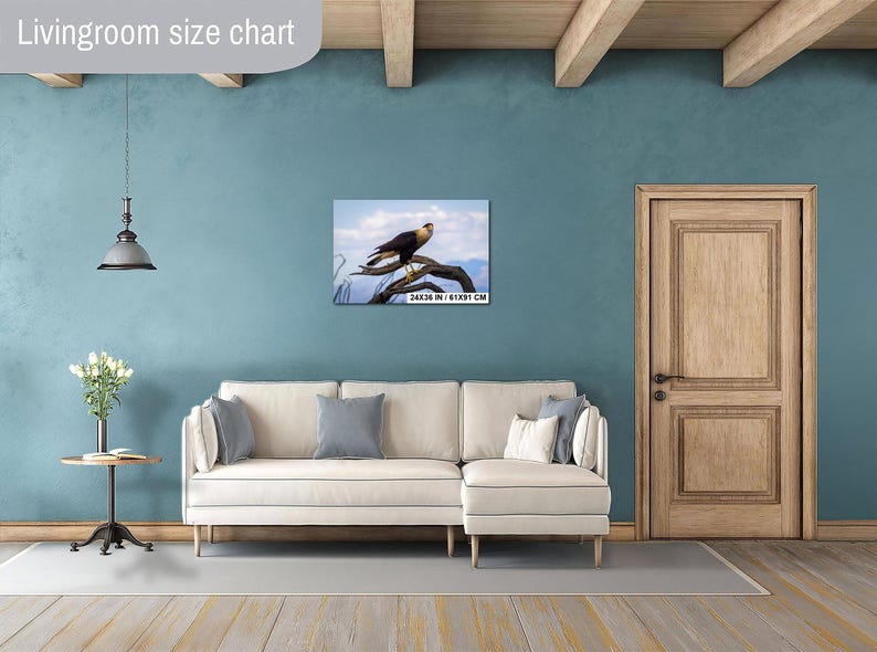 Op de afbeelding: Een ingelijste print van een roofvogel die op een tak zit, tegen een blauwe lucht. Het kunstwerk wordt tentoongesteld boven een cr&egrave;mekleurige bank in een woonkamer. De print meet 61x91 cm.