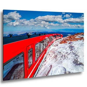 May include: A bright red train car travels along a track on a snowy mountain slope under a vibrant blue sky with white clouds. The train features large rectangular windows and a black roof, contrasting against the rugged, icy terrain and distant peaks.