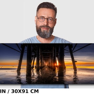 May include: A panoramic photograph of a wooden pier extending into the ocean at sunset. The sun is setting behind the pier, casting a golden glow on the water. The sky is a mix of blue, purple, and orange. The pier is made of dark wood and has many supports. The water is calm and reflects the colors of the sky. The image is 12x36 inches or 30x91 centimeters.