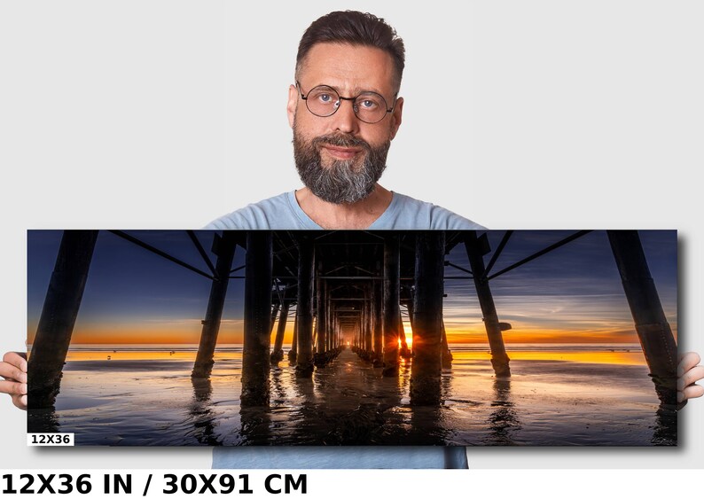 May include: A panoramic photograph of a wooden pier extending into the ocean at sunset. The sun is setting behind the pier, casting a golden glow on the water. The sky is a mix of blue, purple, and orange. The pier is made of dark wood and has many supports. The water is calm and reflects the colors of the sky. The image is 12x36 inches or 30x91 centimeters.
