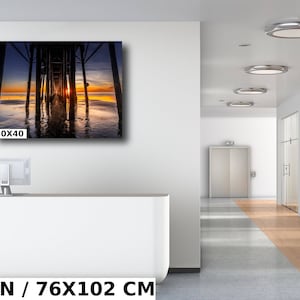 May include: A photograph of a wooden pier extending into the ocean at sunset. The sun is setting behind the pier, casting a warm glow on the water. The pier is made of wooden planks and has a railing on either side. The water is calm and reflects the colors of the sunset. The image is 30x40 inches.