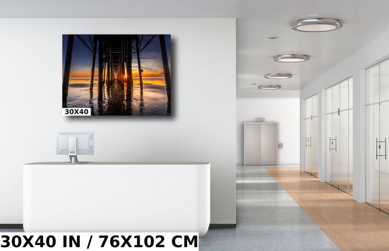 May include: A photograph of a wooden pier extending into the ocean at sunset. The sun is setting behind the pier, casting a warm glow on the water. The pier is made of wooden planks and has a railing on either side. The water is calm and reflects the colors of the sunset. The image is 30x40 inches.