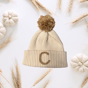 Op de afbeelding: Een crèmekleurige gebreide beanie met een bruine pompon en een bruine geborduurde letter C.