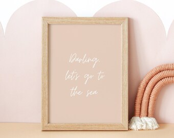 May include: A framed print with the words "Darling, let's go to the sea" in white script. Beside it is a woven rainbow in shades of peach and cream, set against a pink and cream background. The overall theme is nursery decor.