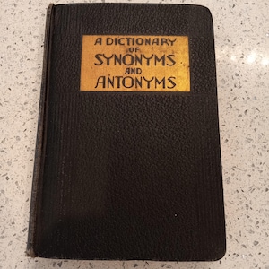 May include: A vintage dictionary of synonyms and antonyms. The book has a black cover with a gold rectangular label that reads "A Dictionary of Synonyms and Antonyms". The book is closed and appears to be well-worn.