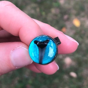 May include: A ring with a blue and black shark tooth design. The tooth is set in a clear resin dome and the ring is made of black metal.