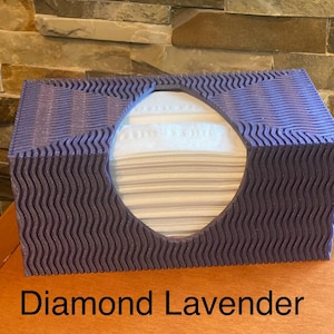May include: A lavender tissue box with a wavy, textured design. The box has a large oval opening in the center, revealing a stack of white tissues. The text "Diamond Lavender" is printed at the bottom.