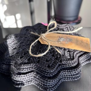 May include: A stack of black, star-shaped crochet coasters tied with twine and a small tag. The coasters have a wavy edge and a textured pattern. The tag has text on it. The coasters are on a dark surface.