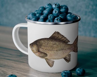 May include: A white enamel mug with a silver rim and handle, featuring a detailed illustration of a fish. The mug is filled with fresh blueberries. The mug is sitting on a wooden surface.
