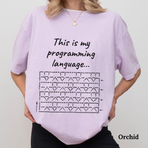 May include: Orchid-coloured t-shirt featuring the text "This is my programming language..." above a black crochet pattern. The shirt has a relaxed fit and short sleeves.