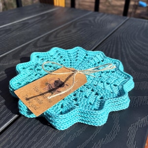Puede incluir: Un conjunto de posavasos de ganchillo turquesa con borde festoneado. Una pequeña etiqueta marrón está atada a los posavasos con hilo, y dice "You make me smile Thank you!" en texto negro. Los posavasos están sobre una superficie gris oscuro.