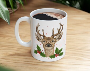 May include: White ceramic mug featuring a smiling deer head with antlers, surrounded by holly and red berries. The mug is filled with dark coffee. A festive holiday-themed design.