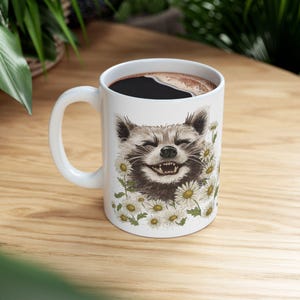 May include: White ceramic coffee mug featuring a smiling raccoon surrounded by daisies. The raccoon has brown and gray fur, and the mug is filled with dark coffee. The mug has a white handle and is set on a wooden surface.
