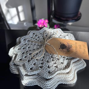 Può includere: Una pila di sottobicchieri all'uncinetto grigio chiaro con un bordo smerlato. Un piccolo tag marrone legato con spago presenta un'illustrazione di un cane e il testo "You make me smile. Thank you!" I sottobicchieri sono impilati su una superficie scura.