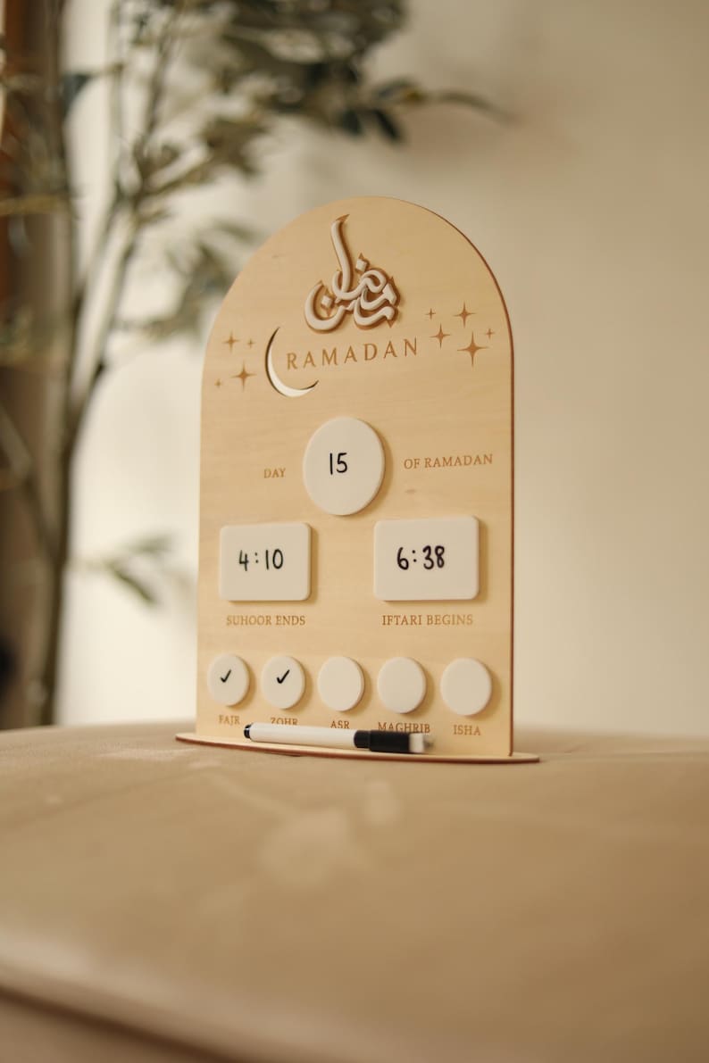 Puede incluir: Calendario de cuenta regresiva de Ramad&aacute;n de madera con la palabra "Ramad&aacute;n" en escritura &aacute;rabe. El calendario muestra el d&iacute;a, las horas de Suhoor e Iftar y las horas de oraci&oacute;n. El d&iacute;a actual es el 15. Incluye un rotulador borrable.
