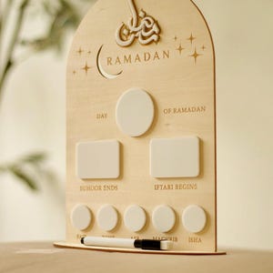 Puede incluir: Calendario de Ramad&aacute;n de madera con dise&ntilde;o de luna creciente y estrellas. El calendario tiene espacios para rastrear eventos diarios, incluidos los horarios de Suhoor e Iftar. Un bol&iacute;grafo blanco descansa en la base.