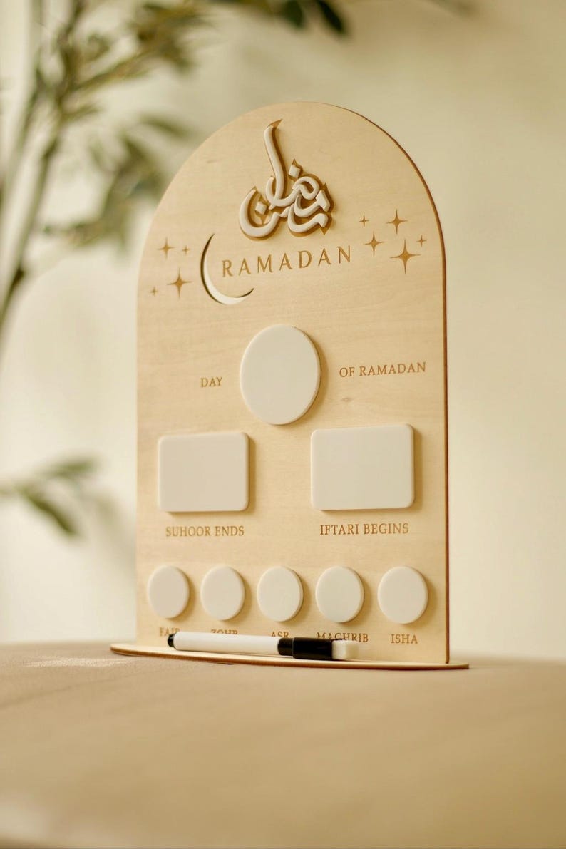 Puede incluir: Calendario de Ramad&aacute;n de madera con dise&ntilde;o de luna creciente y estrellas. El calendario tiene espacios para rastrear eventos diarios, incluidos los horarios de Suhoor e Iftar. Un bol&iacute;grafo blanco descansa en la base.