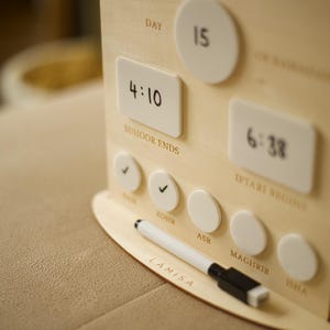 Puede incluir: Calendario de Ramad&aacute;n de madera con marcadores magn&eacute;ticos blancos e indicadores de tiempo. El calendario muestra el d&iacute;a 15, con horarios para Suhoor e Iftar. Un rotulador blanco descansa sobre la base. La palabra "Ramad&aacute;n" est&aacute; arriba.