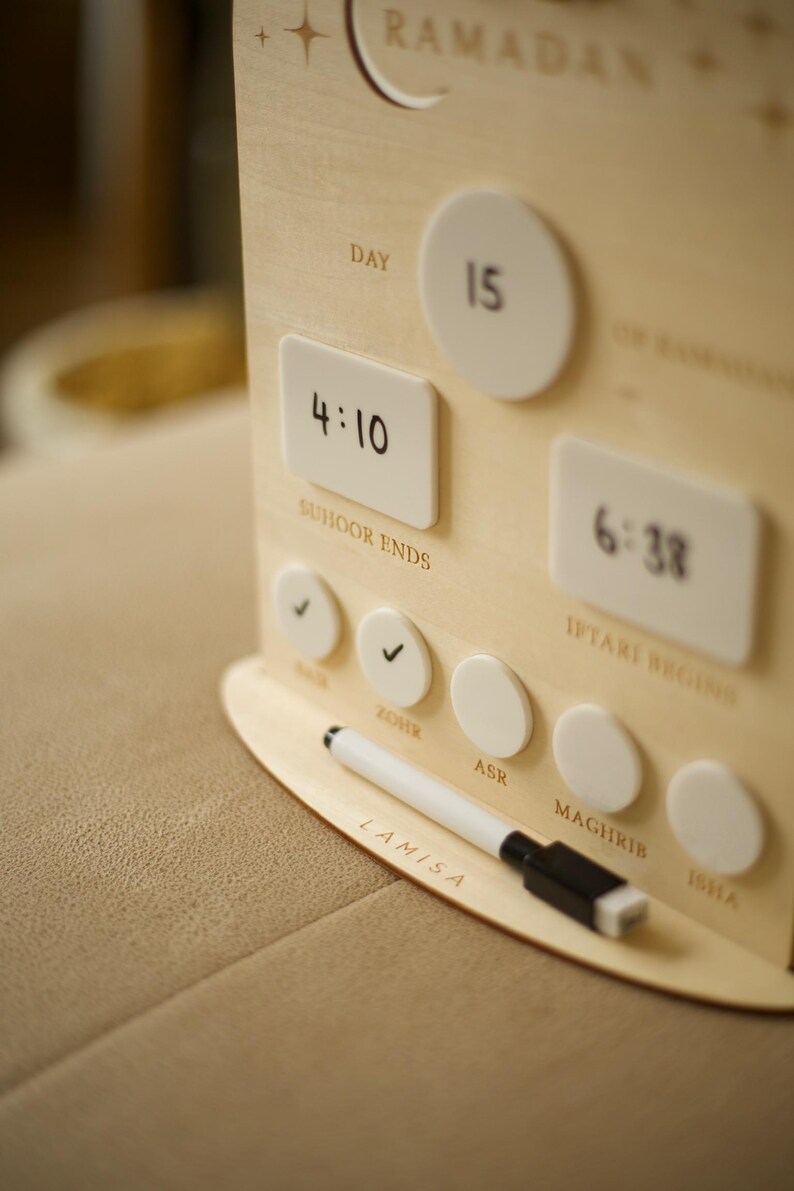 Puede incluir: Calendario de Ramad&aacute;n de madera con marcadores magn&eacute;ticos blancos e indicadores de tiempo. El calendario muestra el d&iacute;a 15, con horarios para Suhoor e Iftar. Un rotulador blanco descansa sobre la base. La palabra "Ramad&aacute;n" est&aacute; arriba.