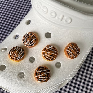 May include: Five brown and white cinnamon roll charms on a white Crocs shoe. The charms are shaped like cinnamon rolls with white icing and brown swirls.