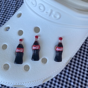 May include: Three miniature Coca-Cola bottle charms in red and black, attached to a white shoe with holes.
