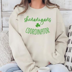 Peut inclure: Un sweat-shirt cr&egrave;me avec le texte "shenanigans coordinator" en lettres vertes avec un tr&egrave;fle vert.