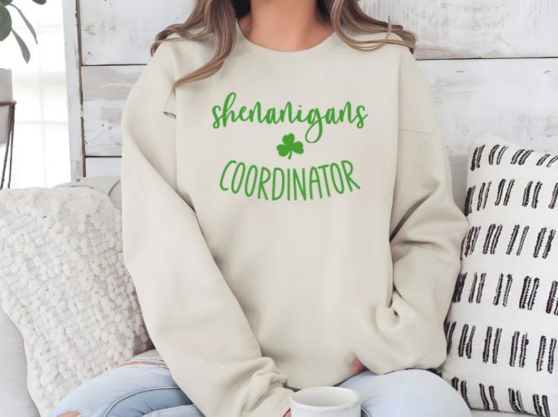 Peut inclure: Un sweat-shirt cr&egrave;me avec le texte "shenanigans coordinator" en lettres vertes avec un tr&egrave;fle vert.