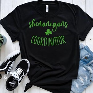 Peut inclure: T-shirt noir avec du texte vert qui dit "Shenanigans Coordinator" et un tr&egrave;fle vert.