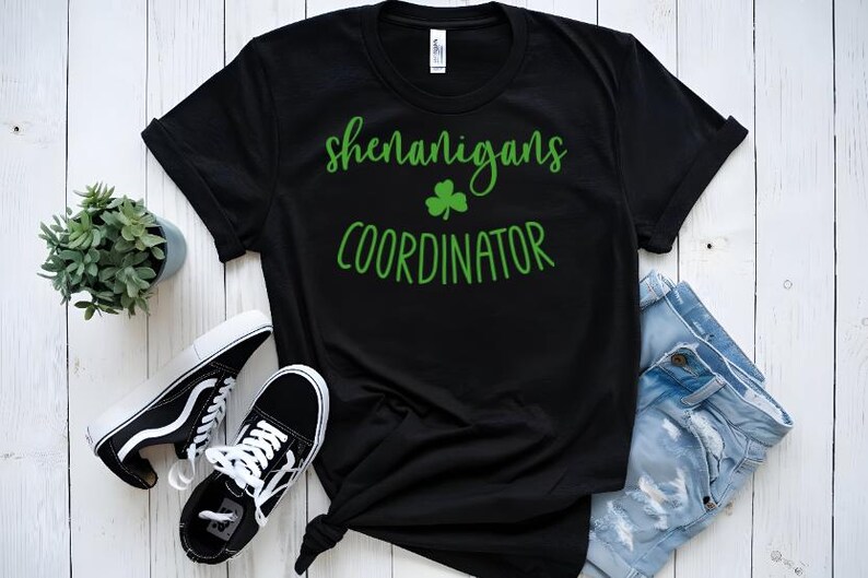 Peut inclure: T-shirt noir avec du texte vert qui dit "Shenanigans Coordinator" et un tr&egrave;fle vert.