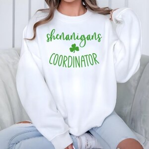 Peut inclure: Sweat-shirt blanc avec du texte vert qui dit "shenanigans coordinator" et un tr&egrave;fle vert.