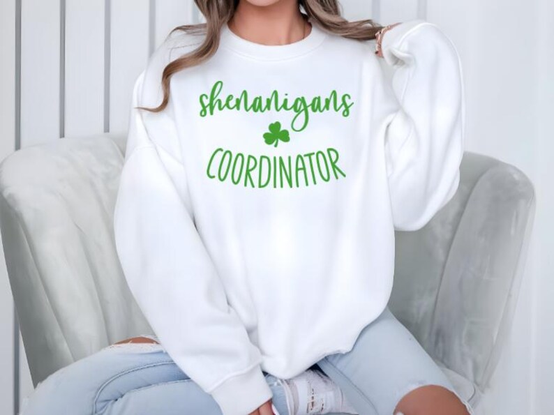 Peut inclure: Sweat-shirt blanc avec du texte vert qui dit "shenanigans coordinator" et un tr&egrave;fle vert.