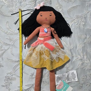 May include: A handmade doll with black yarn hair, a pink flower, and a necklace. The doll wears a pink top, a skirt with a yellow and white lace overlay, and a pink sash. The doll is approximately 20 inches tall.