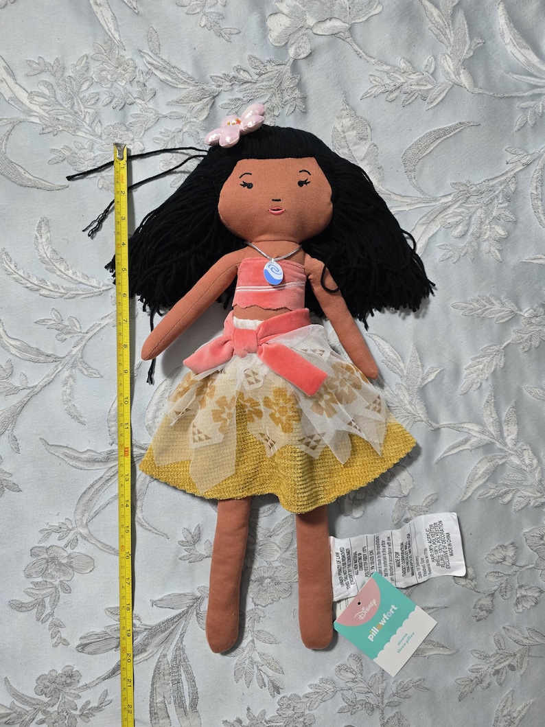 May include: A handmade doll with black yarn hair, a pink flower, and a necklace. The doll wears a pink top, a skirt with a yellow and white lace overlay, and a pink sash. The doll is approximately 20 inches tall.