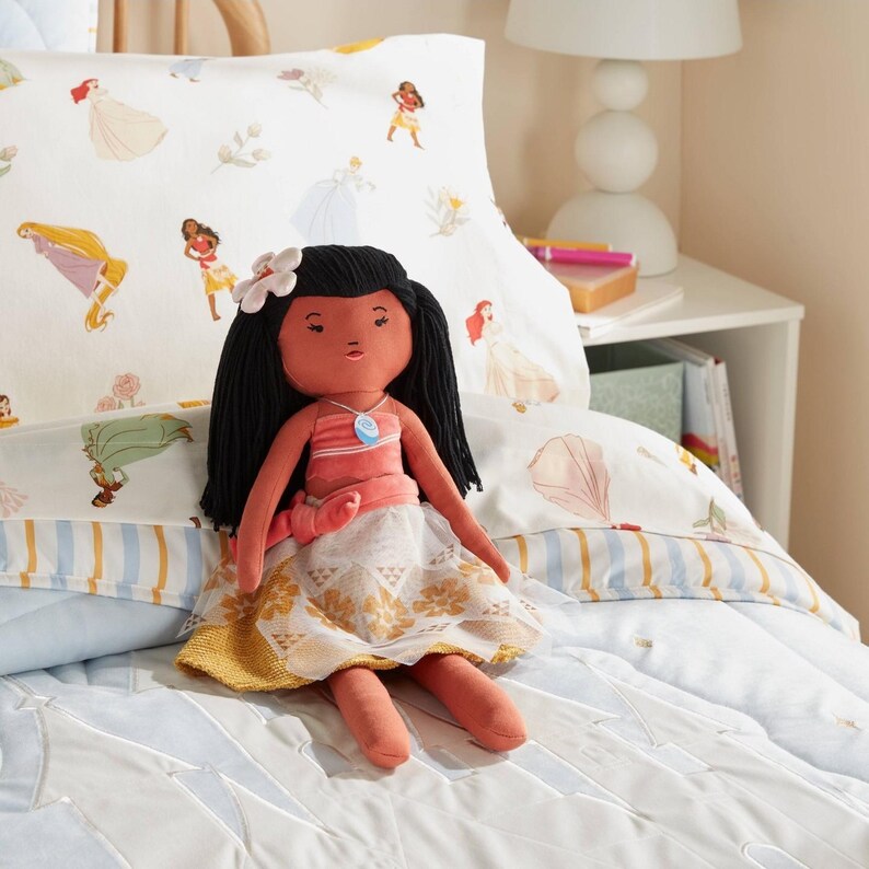 May include: A plush doll with black hair, wearing a coral top, a white skirt with a gold floral pattern, and a flower in her hair. The doll is sitting on a bed with a princess-themed comforter and pillowcase.