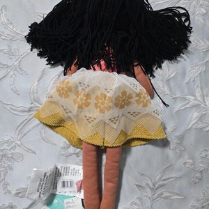 May include: A handmade doll with black yarn hair and a yellow and white floral skirt. The doll has long, slender legs and a pink torso. Tags are attached to the doll.