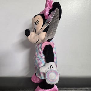 May include: A plush Minnie Mouse doll, featuring a black head, ears, and a pink bow. She wears a plaid dress in pastel colors and pink shoes. The doll has a white glove with a "TRY ME! PRESS HERE" tag.